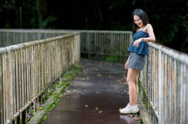 Genç bir kadın yemyeşil bir ormanda dingin bir patikada duruyor, çevresinin tadını çıkarıyor. Güneş ışığı ağaçların arasından süzülüp, barışçıl bir atmosfer yaratır..