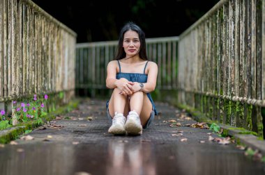 Bereketli bir ormandaki köprüde oturan genç bir kadının huzurlu portresi. Doğa ile çevrili, açık alanının sükuneti ve güzelliğinin tadını çıkarıyor..