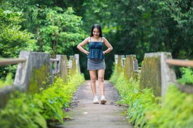 Genç bir kız, yemyeşil bir otlakla çevrili bir orman yolunda huzurlu bir anın tadını çıkarıyor. Bu görüntü doğanın ve sükunetin özünü yakalar. Seyahat ve açık hava temaları için mükemmel.