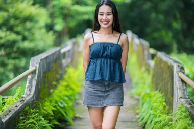 Genç bir kadın yemyeşil bir orman yolunda yürüyor, doğanın güzelliğinin tadını çıkarıyor. Gülümsüyor ve rahatlıyor. Canlı yeşillikler arasında huzur ve sükuneti simgeliyor..