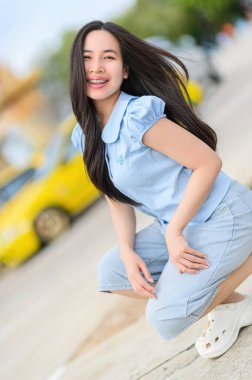 Açık mavi bir elbiseyle dışarıda oturan genç bir kadın neşeyle gülümsüyor. Bu neşeli portre yazın ve şehir hayatının özünü yansıtıyor..