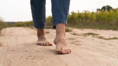 Woman walking in field meadow. Close-up of bare feet soiled with the ground. Female legs on a rural sand road. Healthy lifestyle concept.