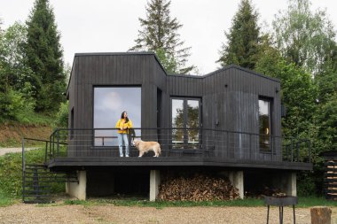 Sarı ceket ve kot pantolon giyen bir kadın ve modern İskandinav tarzı ahşap kulübenin yanında golden retriever köpeği. Yavaş yaşam ve hygge yaşam tarzı konsepti.