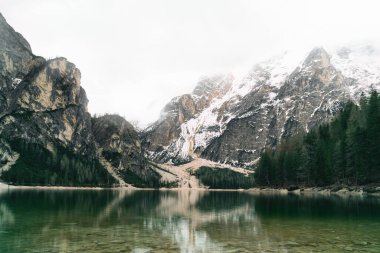 Braies Gölü 'nün panoramik manzarası, Alpler, İtalya. Sakin bir alp gölü üzerinde bulutlu bir gökyüzü beliriyor. Suyun kenarından yükselen dramatik, karla kaplı dağ zirvelerini yansıtıyor..