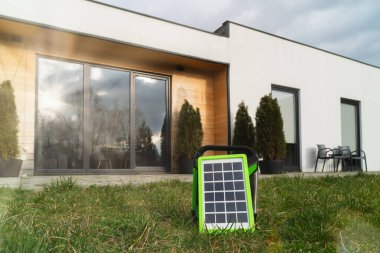 Small solar panel in green frame on lawn, contemporary home with large windows in background. clipart