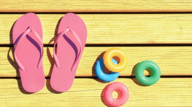 Parlak pembe parmak arası terlikler sarı ahşap döşeme üzerinde renkli havuz yüzen oyuncakların yanında duruyor. Canlı sahne güneşli bir gün yakalıyor, havuz kenarında dinlenmek için mükemmel, ve yaz eğlencesini vurguluyor..