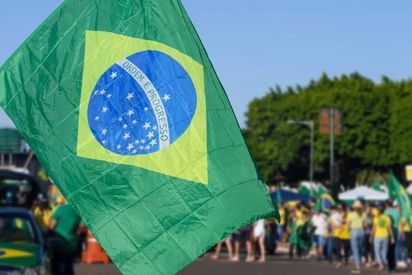 Brazilian flag focused on foreground and brazilian people on the streets blurred on the background. Blurred people wearing green and yellow clothes. Concept of soccer or political background.