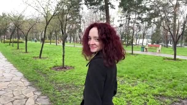 春雨中 一个年轻的女人 留着一头红润的卷发 在公园里散步 微笑着 — 图库视频影像