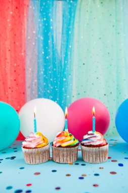 Mavi arka planda mumları olan üç gökkuşağı renkli kek. Hava balonları, dikey kutlama kartı.