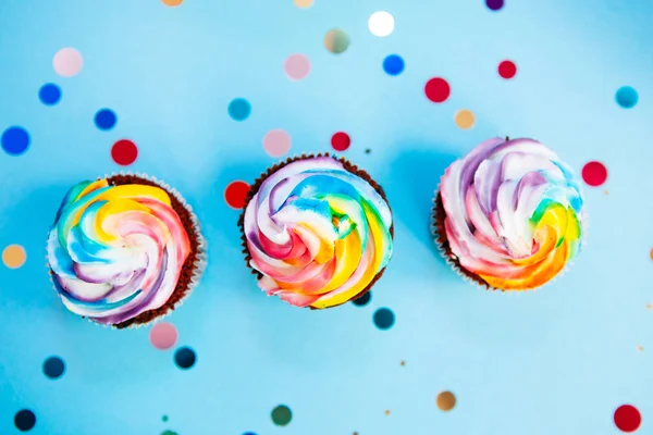 Drei Regenbogenfarben Cupcake Auf Blauem Hintergrund Mit Glitzerkonfetti Von Oben lizenzfreie Stockbilder