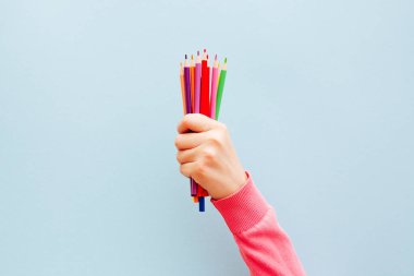 Açık mavi arka planda renkli kalemleri olan kadın eli okula ve sanat eğitimi konsept kartına geri dönüyor.