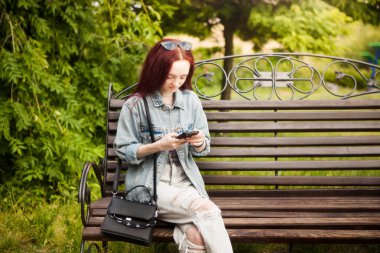 Çilli genç bir kadın bankta oturmuş dışarıda telefonla konuşuyor.