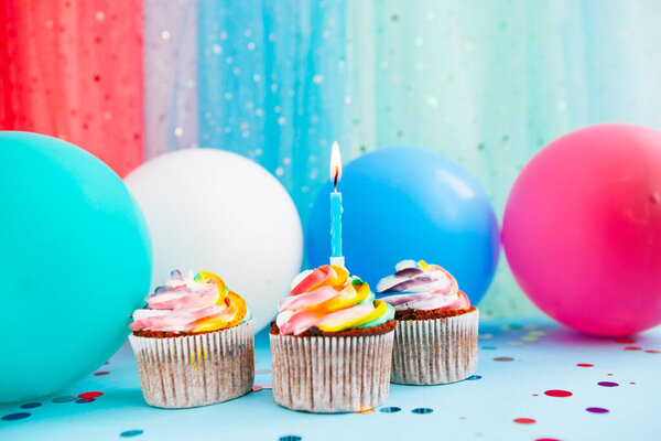 Rainbow colorful muffins or cupcakes with one candle and air balloons on blue, happy birthday card 