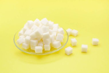 Sarı zemin üzerinde cam tabakta beyaz şeker küpleri, yiyecek malzemeleri.