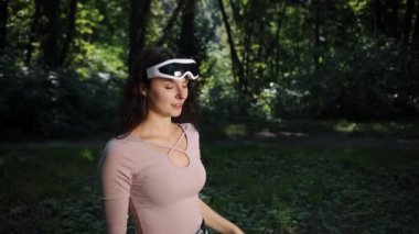 Doğa arka plan, teknoloji ve insan konsepti, VR gözlüklü genç bir kadın gün batımında parkta video oyunları oynuyor doğanın bulanık arka planına karşı.