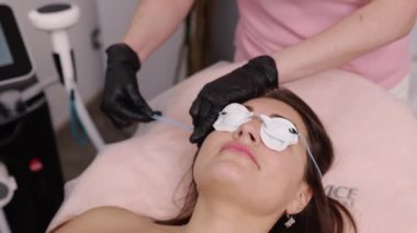 Pamuk pedler, güzellik uzmanı, gözlük takıyor. Kadınların yüzü tıbbi prosedüre tabi tutulur. Elleri eldivenle tutulur. Yüz gençleştirme için lazer verilmeden önce metal göz koruyucu takılır.