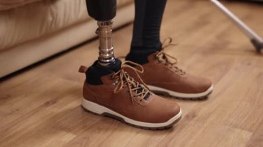 Kanepe oturma, yardımcı cihaz, bağımsız yaşam. Yakın plan gösterileri, protez bacaklı bir adam kanepeye ayakkabılarını giyer, koltuk değneklerini yanında dinlendirir..