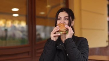 Hamburger keyfi, günlük yemek yeme, restoran çevresi. Esmer saçlı genç bir kadın lokantanın hemen dışında hamburgerle yemek yiyor..