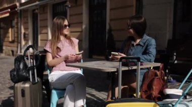 Çift oturuşu, Kadın Turistler, Bekleyen Kadınlar. Seyahatte olan iki kadın, bavullarıyla, telefonlarını kullanırken masada oturuyorlar..