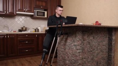 Adaptasyon ekipmanları, yapay uzuv, işyeri ergonomisi. Gözlüklü ve bacak protezli bir adam dizüstü bilgisayarda çalışıyor. Koltuk değnekleri yakınlarda..