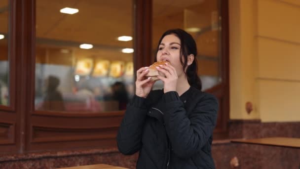 ブルネットバーガー 屋外での食事 若いダイナー 若い黒髪の女性はレストランの近くに屋外でバーガーを楽しんでいます — ストック動画