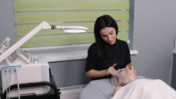 清洁洗液 诊所程序 病人经验 在美容院 熟练的美容师正在进行面部按摩 并在顾客的脸上涂润肤霜 — 图库视频影像