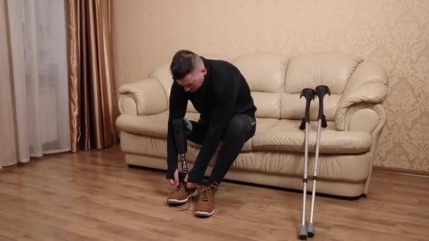 身体障害者 義肢テクノロジー 脚切断 近くに横たわっている彼のクラッチで 障害のために義足を使用する男性はソファーに座って靴を履いています — ストック動画