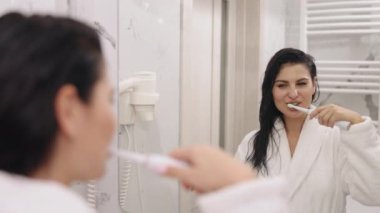 Bornoz, beyaz havlu, kadın bornozu. Güzel bayan, yeni yıkanmış, dişlerini fırçalarken banyo aynasına bakıyor, beyaz, ipeksi bornoz giymiş.