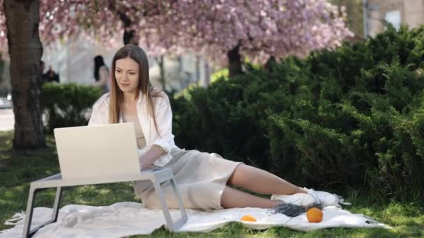 距離学習 アウトドアオフィス 桜の花に囲まれて 女性は公園で彼女のラップトップに座って働きます — ストック動画