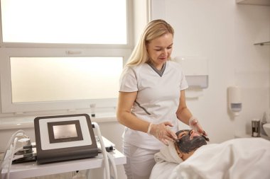 Deri zehirlenmesi, terapi maskesi, spa deneyimi. Profesyonel kaplıcanın parlak atmosferinde, konsantre estetikçi derin temizleyici kömür maskesi uyguluyor, müşterilerin her santimini kaplıyor..