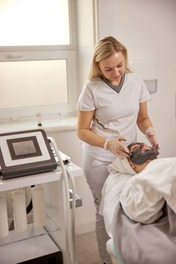 İyi aydınlatılmış spa, güzellik merkezi, cilt tazeleme. Estetisyen, müşterilerin yüzlerini dikkatlice detoksifikasyon yapan kömür maskesiyle kaplıyor, iyi aydınlatılmış spa sınırlarında kapsamlı bir temizlik hedefliyor..