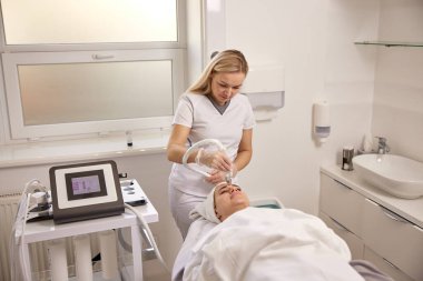 Terapi, güzellik hizmetleri, gençleştirme teknikleri. Güzellik kliniğindeki kendini adamış güzellik uzmanı yüz gençleştirme için ultrason terapisi yapmaya odaklanıyor..