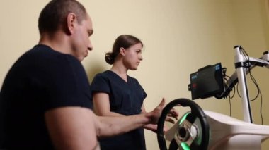 Kol terapisi, el egzersizleri, iyileşme terapisi. Özel rehabilitasyon kliniğinde, hasta kol ve el iyileştirme egzersizlerini yeni bir direksiyon cihazı kullanarak gerçekleştiriyor, terapistler dikkatli gözlerle.