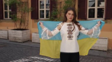 Kendini sarmış, ulusal gurur, Urban arkaplanı. Geleneksel işlemeli bluz giyen genç bir kadın, ahşap ekicilerle birlikte tuhaf kentsel arka planda mavi ve sarı bayrağı gururla açıyor..