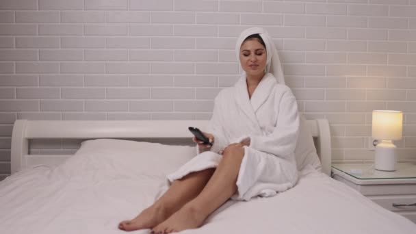 在家里 频道跳跃 休闲活动 穿浴衣的女士正在她的卧室里用遥控器通过电视节目进行转换 — 图库视频影像