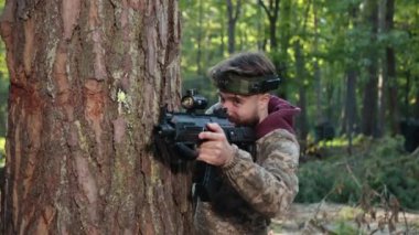 Lazer etiketi, ağaç desteği, taktik teçhizat. Kamuflaj kıyafetli ve kafasında işaretle ağaca yaslanmış bir adam dikkatlice lazer silahı doğrultuyor ve ormanda düşmana ateş ediyor..