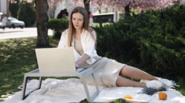 Park 'ta dizüstü bilgisayar, Sakura Parkı, Çiçek Özgeçmişi. Bayan, sakura ağaçlarıyla çevrili olarak parkta dizüstü bilgisayarıyla çalışıyor..
