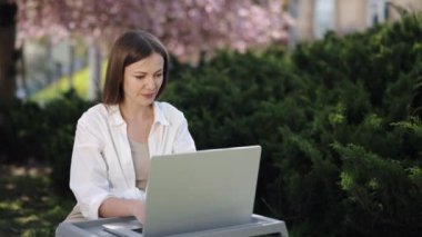 Dijital Göçebe, Kadın Çalışıyor, Sakura Günışığı. Çiçek açan sakura ağaçlarıyla çevrili olan kadın, parktaki dizüstü bilgisayarına odaklanmış durumda..