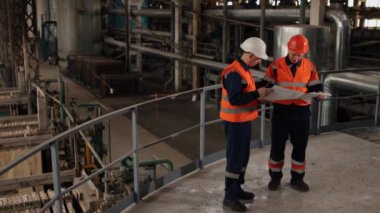 Mühendislik koordinasyonu, maliyet analizi, proje zaman çizelgesi. Fabrika ışıkları, detaylı inşaat tasarımlarını incelerken mühendis ve inşaatçıların üzerine parlıyor..
