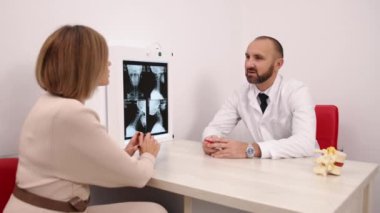 Omurga röntgeni, Sağlık Tartışması, Omurga Sağlığı. Beyaz önlüklü erkek doktor, klinikteki kadın hastayla omurilik röntgeni görüntülerini tartışıyor..
