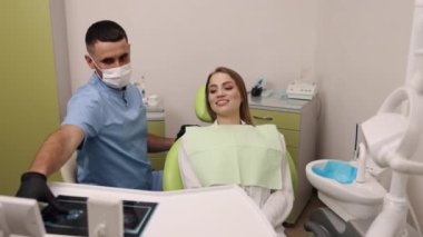 Diş röntgeni, diş muayenehanesi, diş muayenesi. Mavi önlüklü ve maskeli dişçi sandalyedeki gülümseyen hastaya diş röntgeni gösteriyor..