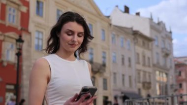 Kadın sohbeti, kadın sohbeti, telefon konuşması. Beyaz elbiseli kadın kulağına telefon tutarken gülümsüyor. Şehir merkezinde tarihi binalarla dikiliyor..