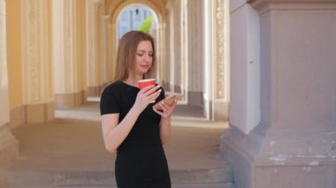 Akıllı telefon kullanmak, akıllı telefon kullanmak, bardağı tutmak. Siyah elbiseli kadın elinde kırmızı kahve fincanı, elinde süslü sütunlarla kemer yolunda duran akıllı telefon kullanıyor..