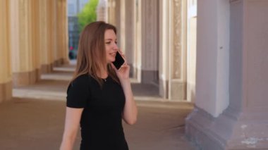 Sohbet eden telefon, şık bir sohbet, şık bir elbise. Siyah elbiseli genç kadın, dekoratif sütunlarla süslenmiş açık hava koridorunda dururken akıllı telefonuyla sohbet ediyor..
