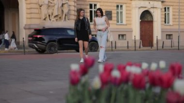 Gezen kadınlar, siyah elbiseler, şehir caddesi. İki kadın şehir caddesinde birlikte yürüyor, biri siyah elbiseli ve ceketli, diğeri beyaz bluzlu ve yırtık pantolonlu, ön planda çiçeklerle.