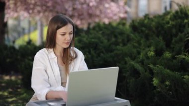 Laptopçu kadın, Blooming Arkaplan, Açık Hava Çalışma Alanı. Sakura 'nın süslediği parkta, kadın dizüstü bilgisayarıyla çalışıyor..