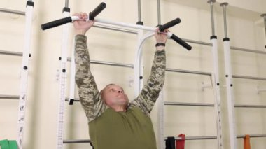 Terapi fitness, kas rehabilitasyonu, adaptasyon eğitimi. Teçhizatla dolu terapi odasında yapay uzuvları olan bir adam duvar parmaklıklarında barfiks egzersizleri yapar..
