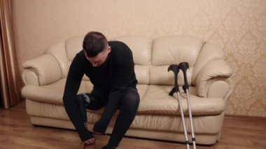 Mobilite yardımı, yardımcı çözümler, denge yardımı. Koltukta oturan, protez bacağı olan bir adam sakatlığı yüzünden koltuk değnekleri yanında dinlenirken ayakkabılarını giyerek giyiniyor..