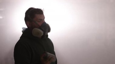 Sprey boyacı, zanaatkar teknikleri, ışıklandırma efektleri. Sis dolu atölyenin ortasında, güvenlik maskeli bir zanaatkar arka plandaki ışıkla vurgulanan sprey boyayla resim yapıyor..