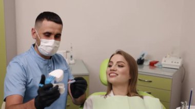 Bekleme modeli, diş konforu, diş bilinci. Diş hekimi, koruyucu yüz maskesi takıyor, dişçi koltuğunda oturan kadın hastaya tedaviyi açıklarken diş modelini ve aletlerini tutuyor.